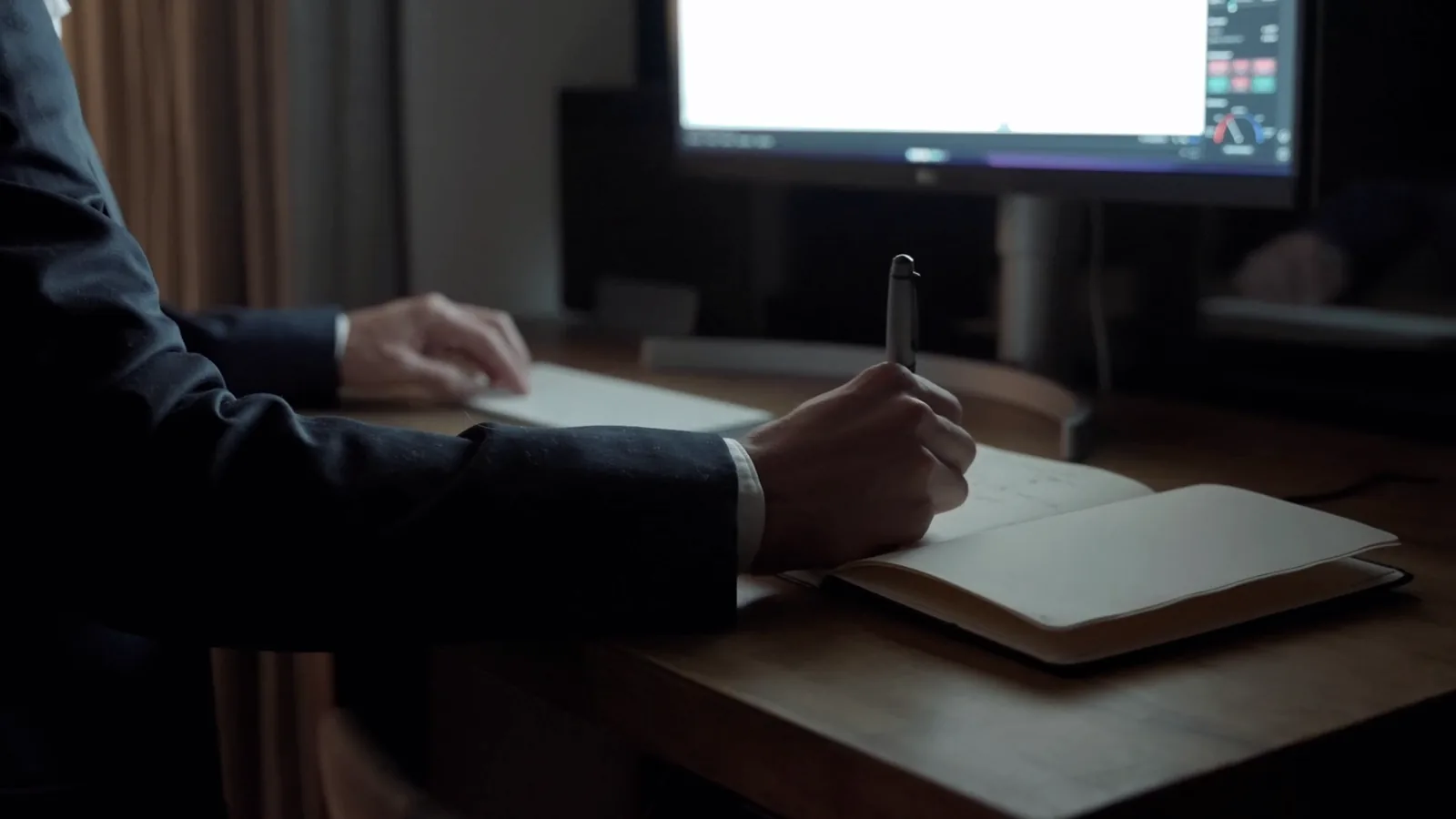 A man in a suit writes in a notebook at a desk, with a trading monitor behind him